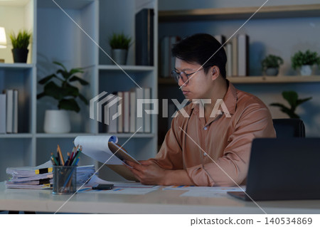 Asian man reviewing paperwork at home office desk late night 140534869