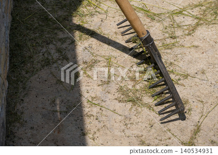 A metal garden rake with a wooden handle rests on dry sandy ground scattered with grass clippings, casting a strong shadow in bright sunlight A metal garden rake with a wooden handle rests on dry sandy ground scattered with grass clippings, casting a strong shadow in bright sunlight 140534931