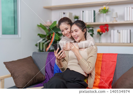 Asian LGBTQ women couple embracing on sofa with pride flag looking at smartphone Asian LGBTQ women couple embracing on sofa with pride flag looking at smartphone 140534974