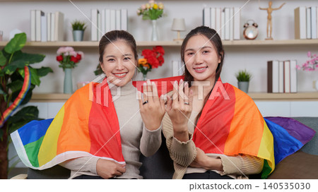 Asian lesbian couple showing engagement rings, wrapped in rainbow pride flag at home Asian lesbian couple showing engagement rings, wrapped in rainbow pride flag at home 140535030