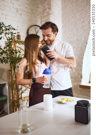 Smiling couple sharing protein drink before workout in bright home kitchen. Smiling couple sharing protein drink before workout in bright home kitchen. 140535350