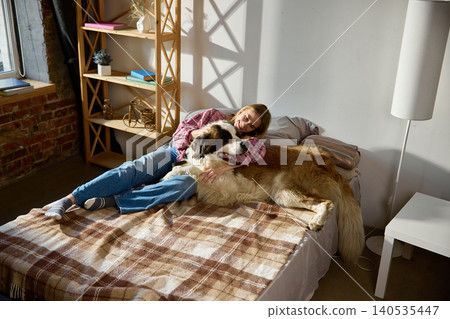 Young woman lying on bed with Saint Bernard dog in sunlit modern apartment interior Young woman lying on bed with Saint Bernard dog in sunlit modern apartment interior 140535447