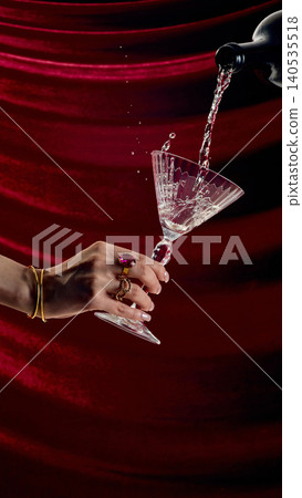 Elegant hand holding crystal martini glass while pouring champagne on red velvet background luxury drink concept Elegant hand holding crystal martini glass while pouring champagne on red velvet background luxury drink concept 140535518