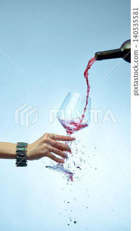 Dynamic splash of red wine pouring into tilted glass with hand on blue background Dynamic splash of red wine pouring into tilted glass with hand on blue background 140535581