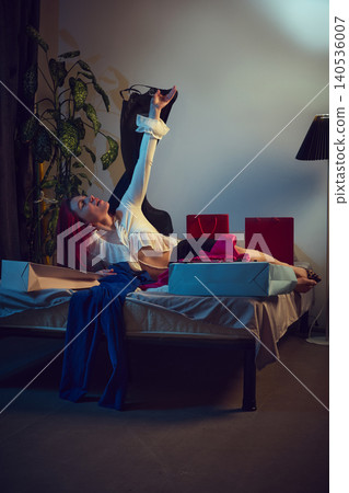 Woman lying on bed trying on clothes from shopping bags at home. Woman lying on bed trying on clothes from shopping bags at home. 140536007