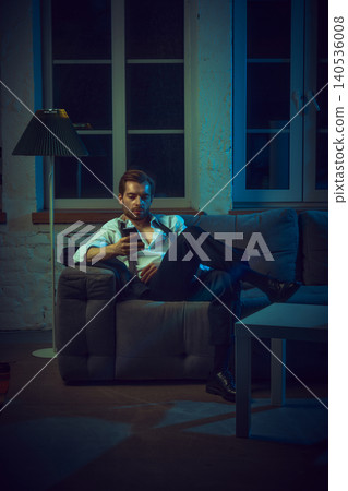 Man sitting on couch checking phone with serious expression in evening interior. Man sitting on couch checking phone with serious expression in evening interior. 140536008