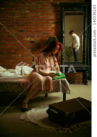 Sad woman sitting on bed holding phone while man reflected in mirror in loft interior. Sad woman sitting on bed holding phone while man reflected in mirror in loft interior. 140536105