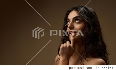 Thoughtful brunette woman touching glossy lips with natural wavy hair on brown background 140536163