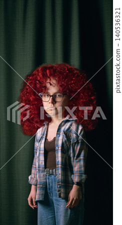 Girl with oversized red curly hair and glasses stands against dark green curtain. Girl with oversized red curly hair and glasses stands against dark green curtain. 140536321