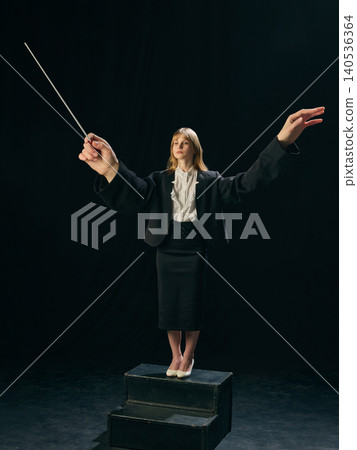 Girl conductor with oversized arms holding baton on dark stage background. Girl conductor with oversized arms holding baton on dark stage background. 140536364