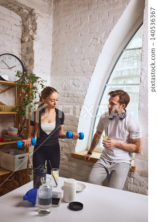 Couple discussing workout routine with dumbbells and drinks in home setting. Couple discussing workout routine with dumbbells and drinks in home setting. 140536367