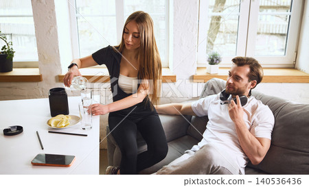 Woman pouring water into glass while man resting on sofa after workout at home. Woman pouring water into glass while man resting on sofa after workout at home. 140536436