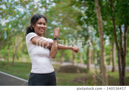 Smiling Black woman stretching arm during outdoor fitness exercise in green park wellness healthy 140536457