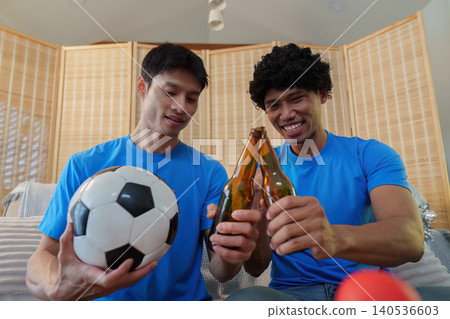 Happy Asian men toasting beer and holding soccer ball at home Happy Asian men toasting beer and holding soccer ball at home 140536603