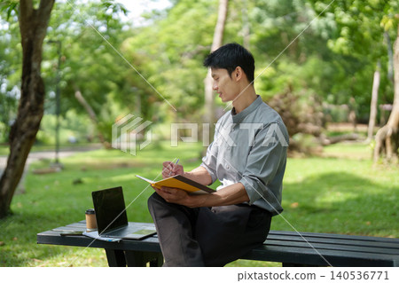 Asian man working and writing outdoors on park bench Asian man working and writing outdoors on park bench 140536771