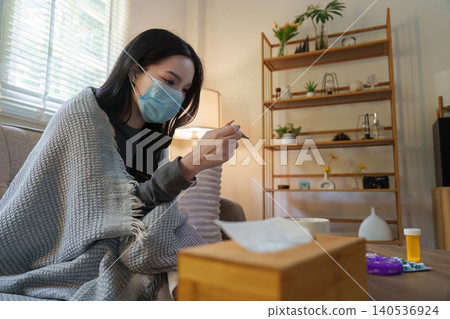 Asian woman with face mask checking temperature with thermometer at home Asian woman with face mask checking temperature with thermometer at home 140536924