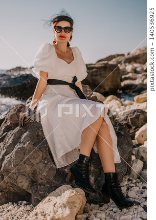 Woman vintage fashion coastal posing on seaside rocks wearing white linen dress and chic accessories during sunny afternoon Woman vintage fashion coastal posing on seaside rocks wearing white linen dress and chic accessories during sunny afternoon 140536925