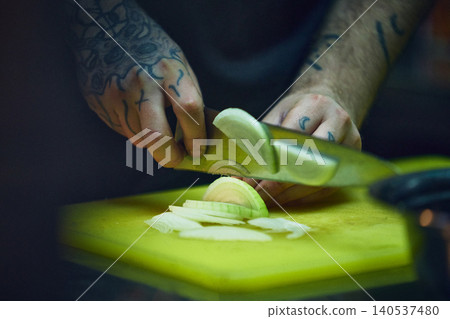 Tattooed chef slicing onion on green cutting board in moody kitchen. 140537480