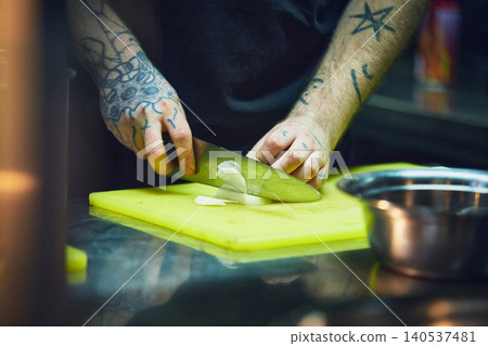Tattooed chef slicing onion on yellow board in busy kitchen. Tattooed chef slicing onion on yellow board in busy kitchen. 140537481