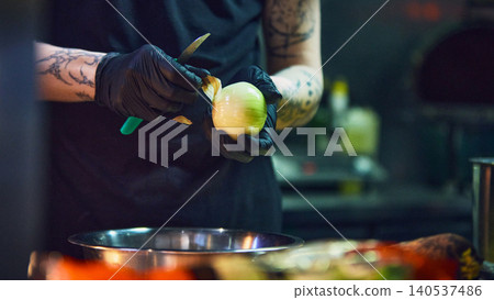 Cook peeling onion with knife over metal bowl in professional kitchen. Cook peeling onion with knife over metal bowl in professional kitchen. 140537486