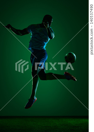 Soccer player midair kicking ball in green dramatic studio light Soccer player midair kicking ball in green dramatic studio light 140537490