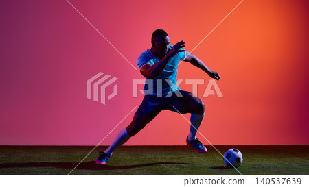 Soccer player sprinting with ball in blue uniform on pink studio field Soccer player sprinting with ball in blue uniform on pink studio field 140537639