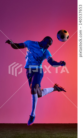 Football player jumping to head ball on grass field against pink orange background. 140537653