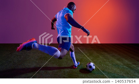 Football player taking powerful kick on grass field with colorful studio lighting. Football player taking powerful kick on grass field with colorful studio lighting. 140537654
