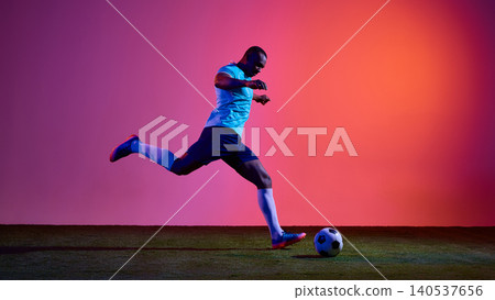 Football player running toward ball on grass field with pink orange studio light. Football player running toward ball on grass field with pink orange studio light. 140537656