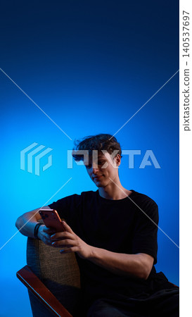 Young man browsing smartphone in chair with blue copy space background. Young man browsing smartphone in chair with blue copy space background. 140537697