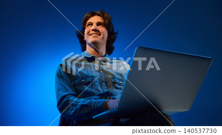 Young man looking upward while using laptop in blue studio light. Young man looking upward while using laptop in blue studio light. 140537730