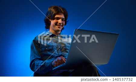 Smiling young man working on laptop against blue gradient studio background. Smiling young man working on laptop against blue gradient studio background. 140537732