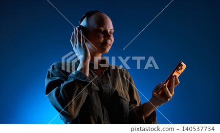 Calm woman listening to headphones while holding smartphone in blue light Calm woman listening to headphones while holding smartphone in blue light 140537734