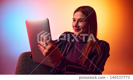 Smiling young woman using tablet on chair against colorful gradient studio background. Smiling young woman using tablet on chair against colorful gradient studio background. 140537773