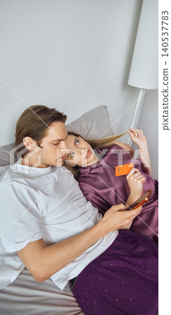 Couple lying in bed choosing online order with phone and orange payment card. Couple lying in bed choosing online order with phone and orange payment card. 140537783