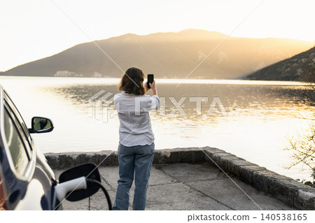 Woman with smartphone next to charging electric car on the seacost during summer vacation. Woman with smartphone next to charging electric car on the seacost during summer vacation. 140538165
