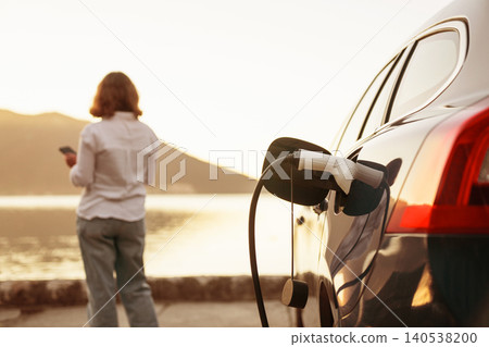 Woman with smartphone next to charging electric car on the seacost during summer vacation. Woman with smartphone next to charging electric car on the seacost during summer vacation. 140538200