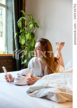 Woman browses smartphone in bed beside LED facial mask in cozy home setting with natural light Woman browses smartphone in bed beside LED facial mask in cozy home setting with natural light 140538702