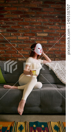 Woman rests on sofa wearing LED facial mask and holding drink, framed by brick wall, soft home light, and cozy interior. Woman rests on sofa wearing LED facial mask and holding drink, framed by brick wall, soft home light, and cozy interior. 140538707