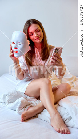 Woman checks smartphone while holding LED facial mask in bed, with soft natural light, white bedding, and clean home setting. Woman checks smartphone while holding LED facial mask in bed, with soft natural light, white bedding, and clean home setting. 140538727