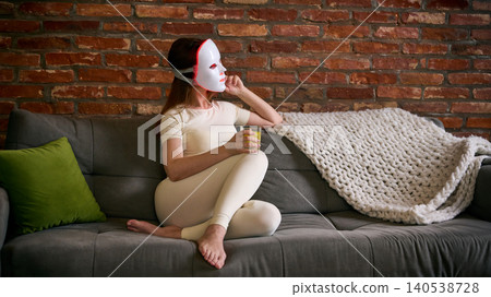 Woman wears LED facial mask on sofa and holds glass of water, with warm home light, brick wall background, and relaxed composition 140538728
