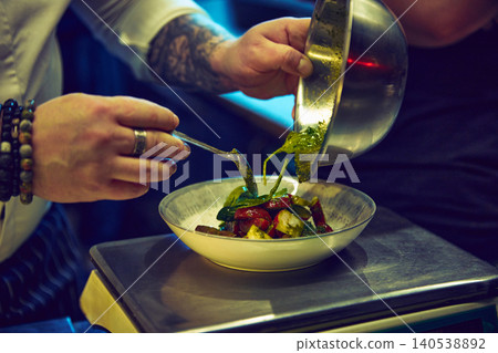 Chef pouring herb sauce over colorful vegetable salad in white ceramic bowl. 140538892
