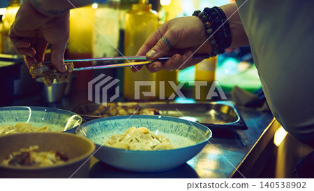 Chef adding roasted meat to creamy pasta bowl on kitchen pass Chef adding roasted meat to creamy pasta bowl on kitchen pass 140538902