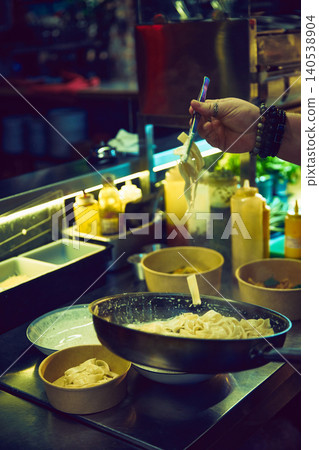 Chef lifting creamy pasta from pan in busy restaurant kitchen. Chef lifting creamy pasta from pan in busy restaurant kitchen. 140538904
