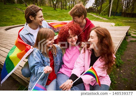 Young friends laughing under rainbow flag on wooden platform in park. 140539377