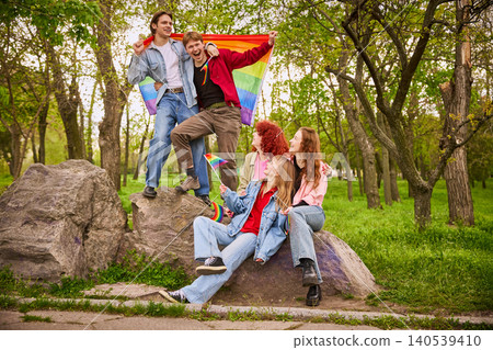 Young friends celebrating with rainbow flag on rocks in sunny park. Young friends celebrating with rainbow flag on rocks in sunny park. 140539410