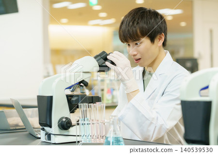一位身穿白色實驗服的日本青年正在使用顯微鏡進行實驗、研究、開發和學習。 一位身穿白色實驗服的日本青年正在使用顯微鏡進行實驗、研究、開發和學習。 140539530
