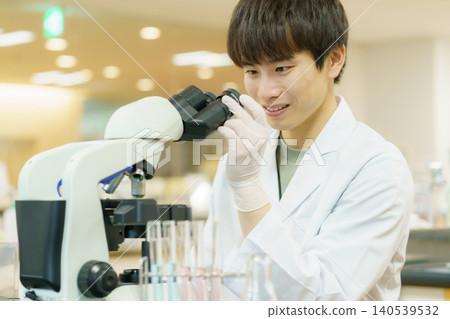 一位身穿白色實驗服的日本青年正在使用顯微鏡進行實驗、研究、開發和學習。 一位身穿白色實驗服的日本青年正在使用顯微鏡進行實驗、研究、開發和學習。 140539532