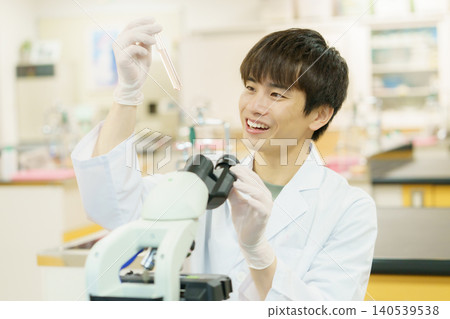 A young Japanese man in a white lab coat working with a microscope. Experiment, research, development, studying. A young Japanese man in a white lab coat working with a microscope. Experiment, research, development, studying. 140539538