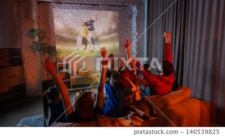 Family celebrating goal while watching soccer game projection in cozy living room. Family celebrating goal while watching soccer game projection in cozy living room. 140539825